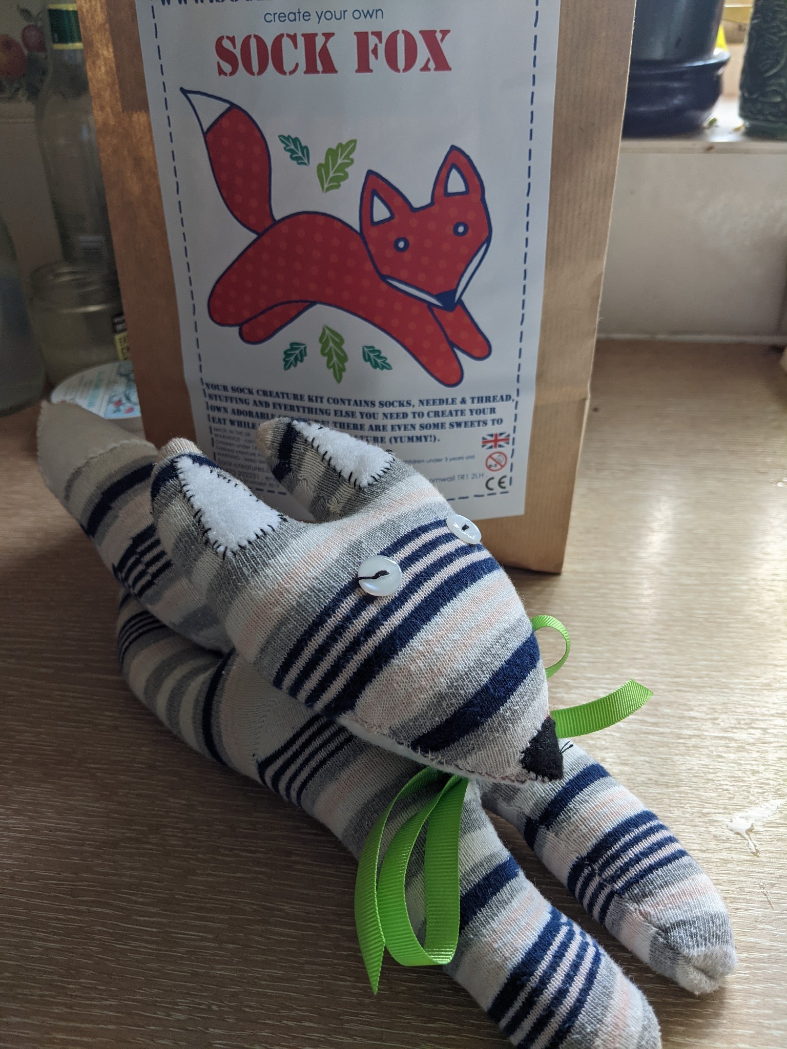 A white, pink and blue striped soft toy fox with button eyes and a green ribbon around its neck.